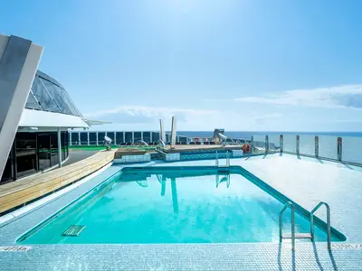 Ein grosser Aussenpool an Bord der MSC Divina unter klarem, blauem Himmel. Der Pool ist von blauen Mosaikfliesen und einem Holzdeck umgeben, dahinter ist das weite Meer zu sehen.