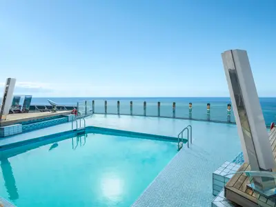 Der The Garden Pool an Bord der MSC Divina: azurblauer Pool auf dem Deck, umgeben von Fliesen und Holz. Weites Meer und klarer blauer Himmel. Moderne, spiegelnde Saeulen ragen empor.