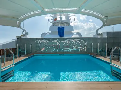 Der hellblaue Pool "The One Pool" im MSC Yacht Club der MSC Divina, umgeben von Holzdeck und einer Wand mit Kunst. Im Hintergrund die Schiffsbrücke. Blauer Himmel mit Wolken.