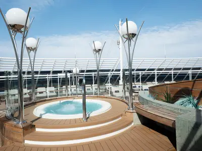 Ein sprudelnder Whirlpool auf einem Holzdeck mit modernen Laternen an Bord der MSC Divina. Dahinter bequeme Liegestühle und eine weiße Überdachung unter blauem Himmel.