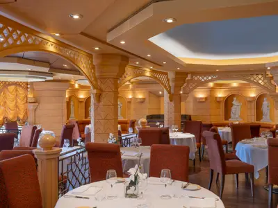 Elegantes Restaurant "Le Muse" an Bord der MSC Divina. Innenraum mit klassischer Architektur: Bögen, Säulen und Statuen in Nischen. Gedeckte Tische mit weißen Tischdecken und rot-braunen Stühlen. Warme Beleuchtung, ohne Personen.