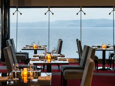 Elegantes Restaurant an Bord der MSC Divina. Gedeckte Tische mit Kerzenlicht und Meerblick durch große Fenster. Graue Stühle, roter Teppich, gemütliche Atmosphäre.