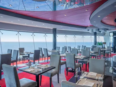 Ein elegantes Spezialitätenrestaurant Galaxy an Bord der MSC Divina mit modernen Tischen und grauen Stühlen, gedeckt für Gäste. Durch große Fensterfronten bietet sich ein weiter Blick auf das offene Meer. Der Raum ist mit einem roten Teppich und stilvoller Deckenbeleuchtung gestaltet.