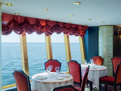 Elegant gedeckte Tische mit roten Samtstühlen im Restaurant Villa Rossa der MSC Divina. Große Fenster bieten weiten Meerblick. Rote Raffrollos und goldene Wanddekorationen prägen das luxuriöse Ambiente.