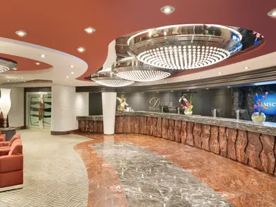 Eine moderne Rezeption an Bord der MSC Divina mit einem langen, geschwungenen Tresen aus dunklem Holz und Marmor. Darüber hängen große, runde Kronleuchter. Links befinden sich rote Sessel.