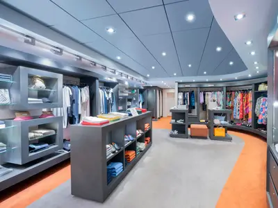 Boutique an Bord der MSC Divina, mit Kleidung, Hüten, Taschen und Schuhen auf Regalen und Kleiderständern. Boden in Grau und Orange, moderne Deckenbeleuchtung.