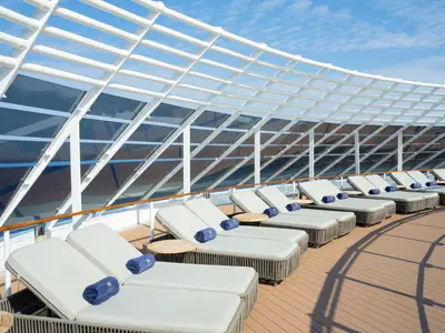 Eine Reihe von leeren Sonnenliegen mit blauen Handtüchern auf einem Holzdeck an Bord der MSC Divina. Darüber wölbt sich eine moderne Glas- und Stahlkonstruktion, die Schatten spendet, während ein strahlend blauer Himmel im Hintergrund zu sehen ist.