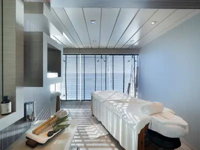 Ein Doppel-Behandlungsraum im MSC Aurea Spa an Bord der MSC Divina. Zwei Massageliegen stehen vor großen Fenstern mit Jalousien, die Meerblick bieten. Pflegeprodukte und Handtücher sind bereitgestellt.