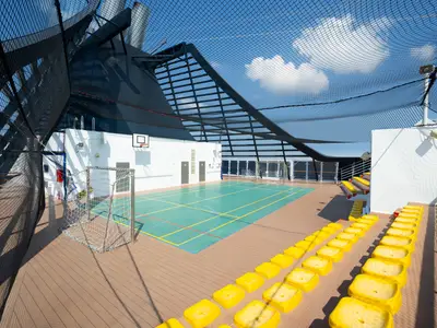 Eine helle Sportarena an Bord der MSC Divina unter freiem Himmel, umgeben von Fangnetzen. Auf dem grünen Multifunktionsfeld sind Linien für Basketball und Fußball markiert. Gelbe Sitze bieten Platz für Zuschauer.