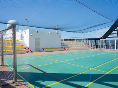 Multifunktions-Sportfeld auf dem Deck der MSC Divina. Türkisfarbenes Spielfeld, gelbe Tribünen, Schutznetz. Bei hellem Tageslicht unter blauem Himmel.
