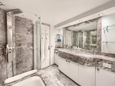 Das luxuriöse Badezimmer einer Suite an Bord der MSC Divina. Es zeigt eine moderne Duschkabine, einen Doppelwaschtisch aus Marmor und Pflegeprodukte. Weiße Bademäntel hängen an der Tür, das Licht kommt von Einbauspots.