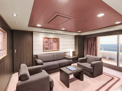 Eine geräumige, elegant eingerichtete MSC Yacht Club Executive & Family Suite an Bord der MSC Divina mit grauer Couch, zwei Sesseln und einem dunklen Couchtisch. Große Fensterfront mit Blick auf das Meer und den privaten Balkon. Die Decke ist teilweise rosa.