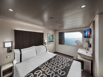 Helle Aussenkabine an Bord der MSC Euribia mit Doppelbett und Nachttischen. Rechts befinden sich ein Schreibtisch mit TV und ein Fenster, das einen weiten Meerblick bietet.