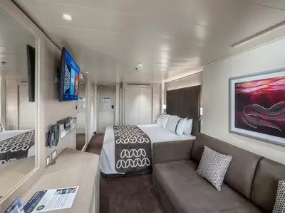 Eine helle und geräumige Balkonkabine an Bord der MSC Euribia, ausgestattet mit einem großen Doppelbett, einem bequemen Sofa, einem Flachbildfernseher und einer eleganten Einrichtung in Grautönen.