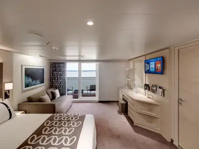 Eine moderne Balkonkabine an Bord der MSC Euribia mit Doppelbett, Sofa und einem Schreibtisch mit TV. Der Balkon bietet zwei Stühle und weiten Meerblick.