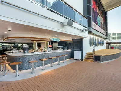 Die Atmosphere Bar North an Bord der MSC Euribia zeigt einen Bartresen mit Hockern. Rechts daneben eine Bühne und eine große LED-Leinwand auf dem Außendeck bei Tageslicht.