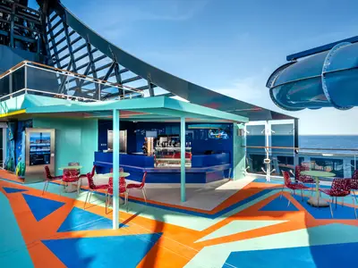 Farbenfrohe Ocean Cay Bar an Bord der MSC Euribia. Modernes Deck mit blauen, orangen Mustern, rotem Gestühl, blauer Bar und Wasserrutsche. Blick aufs Meer.