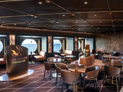 Das Jean-Philippe Chocolat & Café an Bord der MSC Euribia. Dunkle, elegante Einrichtung mit runden Bullaugen-Fenstern mit Meerblick. Braune Sitzbänke und Stühle, sowie Tische mit Glasplatten prägen den Raum.