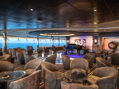 Geräumige Sky Lounge der MSC Euribia mit zahlreichen bequemen Sitzen und Tischen, einer Pianobühne und großen Fenstern mit weitem Blick auf das offene Meer.