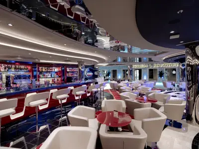 Die elegante Euribia Bar & Lounge an Bord der MSC Euribia. Rote Bartheke, Flaschenregale, weisse Hocker, Sessel und Tische. Glänzende Säulen, Boutique im Hintergrund.