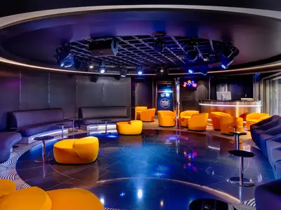 Der moderne und lebhafte Attic Club an Bord der MSC Euribia mit gelben Sitzpoefs, blauen Sofas, einer Bar und Bühnenbeleuchtung für abendliche Unterhaltung.