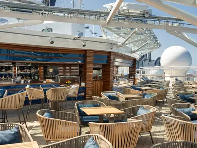 Eine elegante Outdoor-Bar mit Grill und vielen Sitzgelegenheiten an Deck der MSC Euribia unter Tageslicht. Blaue Polster und helle Korbmöbel prägen die Szene.