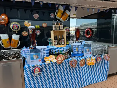 Ein festlich geschmückter Oktoberfest-Biertresen an Bord der MSC Euribia mit Paulaner-Bieren, Brezel-Deko und bayerischen Rautenmustern.