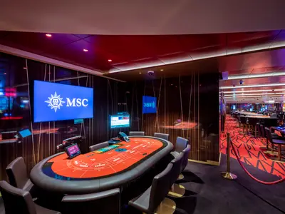 Casino an Bord der MSC Euribia mit rotem Baccarat-Tisch. Dahinter Spielautomaten und weitere Tische auf rotem Teppich. MSC-Logo auf Bildschirm.