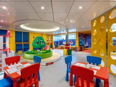 Kinderbereich an Bord der MSC Euribia. Bunte Tische und Stühle, eine grüne Baum-Skulptur mit Lego-Figuren, Modelleisenbahn. Gelbe Wände mit Lochmuster.