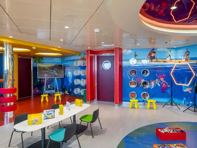 Ein bunter Kinderbereich der MSC Euribia mit Spielzeug, leuchtenden Wandmalereien, einem Basteltisch und farbigen Hockern. Ein glänzender Pfeiler und eine rote Tür sind zu sehen.