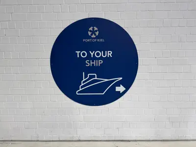 Ein blaues, rundes Hinweisschild an einer weißen Klinkerwand im Hafen von Kiel weist den Weg zur MSC Euribia.