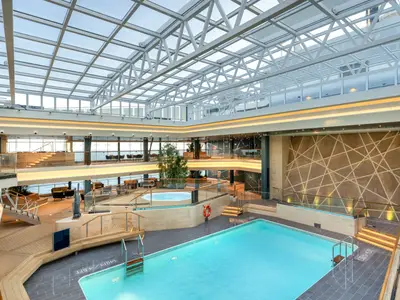 Der weitläufige Bamboo-Pool-Bereich an Bord der MSC Euribia zeigt einen grossen Pool, einen Whirlpool und mehrere Ebenen. Tageslicht fällt durch das Glasdach und eine moderne Wandgestaltung prägt den Raum. Ohne Personen.