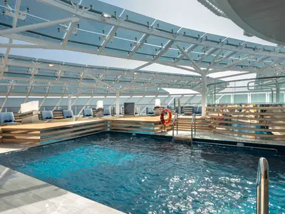 Ein weitwinkeliger Blick auf den eleganten, überdachten Pool an Bord der MSC Euribia. Türkisfarbenes Wasser reflektiert das Tageslicht, umgeben von Holzdecks und bequemen blauen Liegen. Eine moderne Glasdachkonstruktion lässt viel Licht herein.