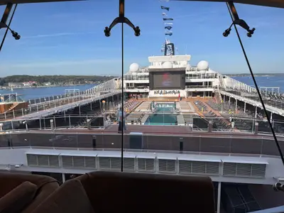 Blick durch eine Glasscheibe auf das weitläufige, sonnige Hauptpooldeck an Bord der MSC Euribia mit Liegestühlen und großem Bildschirm.
