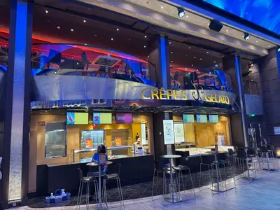 Blick auf den beleuchteten Jean-Philippe Crêpes & Gelato Stand an Bord der MSC Euribia mit Tischen und Stühlen davor.