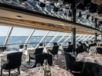 Elegantes Restaurant der MSC Euribia mit weiss eingedeckten Tischen, schwarzen Stühlen und grossen Panoramafenstern mit Meerblick. Eine glänzende Decke reflektiert den Raum.