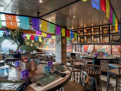 Das mexikanische ¡HOLA! Tacos & Cantina an Bord der MSC Euribia. Bunte Deko, Teller und Gläser auf Holztischen und ein rundes Fenster mit Meerblick sind zu sehen.