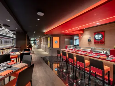 Die stylische Kaito Sushi Bar an Bord der MSC Euribia. Man sieht eine lange Theke mit roten Hockern und Esstische, daneben eine Promenade mit kunstvollen Bögen. Dominierende Farben sind Rot, Schwarz und helle Holztöne.