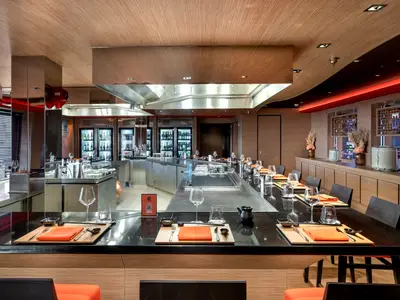 Ein modernes Teppanyaki-Restaurant an Bord der MSC Euribia. Es zeigt eine lange Theke mit Kochflächen, vorbereiteten Gedecken und Sitzgelegenheiten. Im Hintergrund sind Getränkekühlschränke zu sehen.