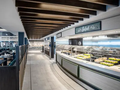 Modernes Marketplace Buffet der MSC Euribia. Eine lange Salatbar mit frischen Speisen in grünen Schalen ist im Vordergrund. Weitere Buffets und geräumige Sitzbereiche im Hintergrund.