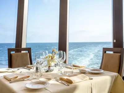 Ein gedeckter Tisch im Spezialitätenrestaurant der MSC Euribia mit Blick auf das offene Meer. Gläser, Besteck und Blumen schmücken den Tisch. Eine elegante und ruhige Szene.