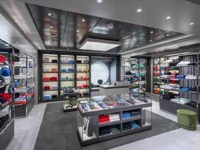 Moderner Bekleidungsshop an Bord der MSC Euribia. Farbige Kleidung, Schuhe und Accessoires sind in beleuchteten Regalen und Vitrinen ausgestellt. Ein rundes Fenster im Hintergrund.