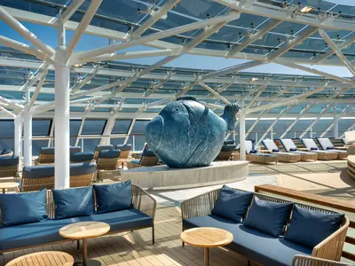 Weitwinkelansicht des luxuriösen Sonnendecks mit großem blauen Fisch als Skulptur an Bord der MSC Euribia. Bequeme blaue Lounge-Möbel unter einer transparenten Glasdachkonstruktion laden zum Entspannen ein, während im Hintergrund das offene Meer zu sehen ist.