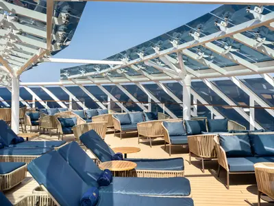 Blick auf das Sonnendeck der MSC Euribia. Blaue Liegen und Sofas mit Rattan-Optik stehen unter einer eleganten Glasüberdachung. Das Meer ist im Hintergrund sichtbar.