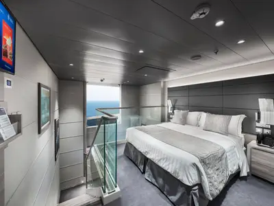 Luxuriöse Duplex Suite an Bord der MSC Euribia. Großes Doppelbett, eleganter Nachttisch und Glasgeländer. Panoramafenster mit weitem Meerblick. TV und Kunst an der Wand.