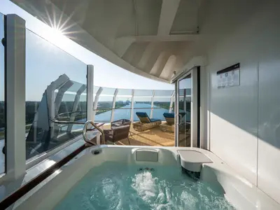 MSC Euribia: Luxus-Suite mit Whirlpool auf dem Balkon. Panorama-Glasfronten bieten weiten Blick auf Fluss, Schiffe und Industrie am Horizont.
