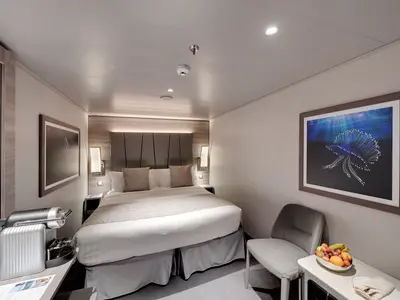 Eine helle und moderne MSC Yacht Club Interior Suite an Bord der MSC Euribia mit Doppelbett, Nachttischen, Sessel, Kaffeemaschine und Obstschale.