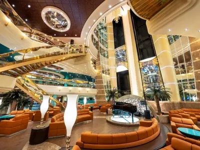 Blick auf das mehrstöckige Atrium der MSC Fantasia mit glänzenden goldenen Treppen, Glasaufzügen, einer Bühne mit Flügel und bequemen orangefarbenen Sitzbereichen.