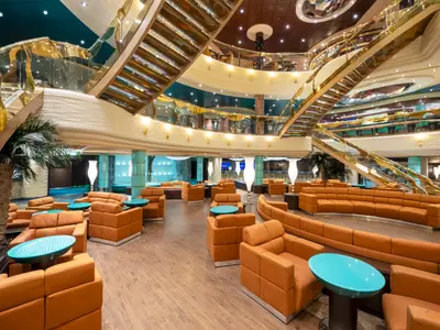 Das opulente Atrium der MSC Fantasia zeigt eine mehrstöckige Lounge mit orangefarbenen Sofas und Sesseln, hellblauen Tischen, zwei glänzenden goldenen Treppen und hohen Decken. Grüne Palmen schmücken den Raum.