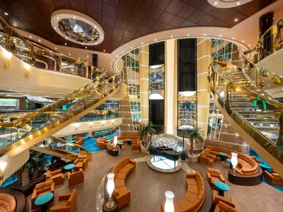 Das luxuriöse Atrium der MSC Fantasia erstreckt sich über mehrere Ebenen mit breiten goldenen Treppen, gläsernen Panoramaaufzügen, bequemen orangefarbenen Sitzgruppen und einem Flügel auf einer beleuchteten Plattform.